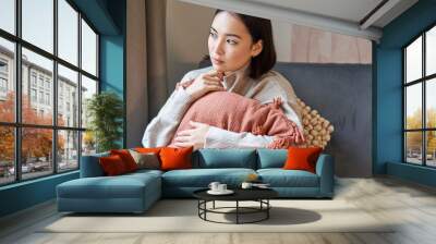 Portrait of asian woman feeling cozy in her home, hugging pillow on sofa and looking thoughtful, smiling Wall mural