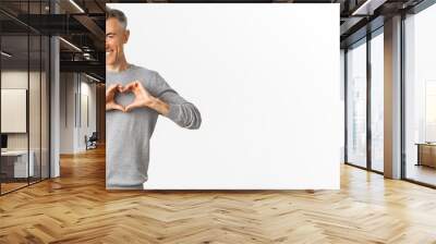 Portrait of cheeky middle-aged man in grey sweater, smiling and showing heart sign, love someone, standing over white background Wall mural