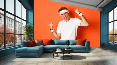 Portrait of confident and cheerful middle-aged man in sport clothing, raising hands up and triumphing, achieve workout goal, standing pleased over orange background Wall mural