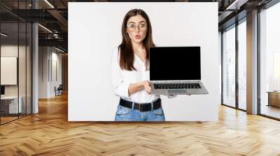 Portrait of cute girl in glasses, student showing laptop screen with amazed face, standing over white background Wall mural