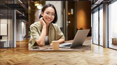 Portrait of smiling girl in glasses, sitting with laptop in outdoor cafe, drinking coffee and working remotely, studying online Wall mural