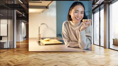 Portrait of smiling young happy woman staying at home, standing in kitchen and eating toast, looking aside Wall mural