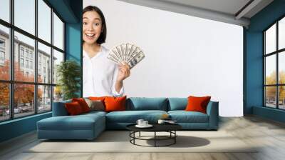 Portrait of young asian woman pointing at her money dollars, showing cash, standing over white background Wall mural