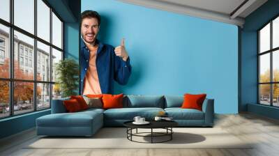 Positive smiling guy showing thumbs up and smiling, complimenting you, praising good job, well done gesture, standing on blue background Wall mural