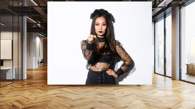 Sassy elegant young asian woman in witch halloween costume tell come closer, flicking finger as lure you, standing over white background Wall mural