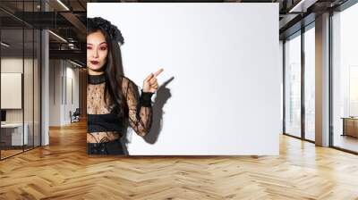 Sassy young evil witch with gothic makeup and wreath, looking arrogant while pointing fingers sideways, showing two halloween themed banners, standing over white background Wall mural