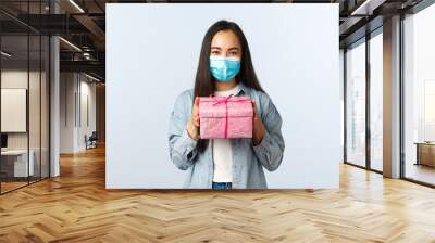 Social distancing lifestyle, covid-19 pandemic, celebrating holidays during coronavirus concept. Cute asian girl in medical mask prepared present for friends birthday, give pink gift box Wall mural