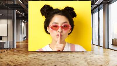 Summer fashion concept. Close-up portrait of glamour asian woman in sunglasses telling a secret, asking be quiet, making hush taboo sign and winking at camera Wall mural