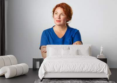 Thoughtful redhead nurse, physician or female doctor in scrubs looking upper left corner with intrigued, interested expression, cross arms chest, turn attention to banner, grey background Wall mural