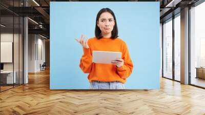 Woman with digital tablet, looks confused, cant understand smth on her gadget, stands puzzled against blue background Wall mural