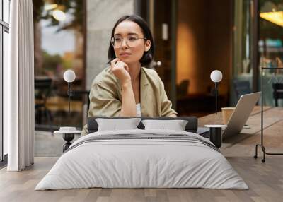 Working woman sitting with laptop in cafe and thinking. Asian girl in glasses works remotely, drinks coffee and looks thoughtful Wall mural