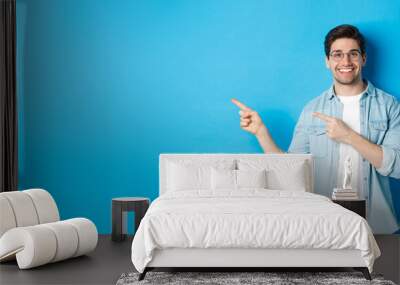 Young handsome man in glasses showing advertisement, smiling and pointing fingers left, making announcement, standing against blue background Wall mural