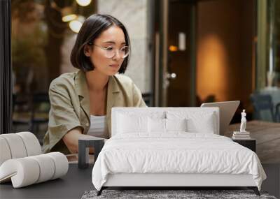 Young woman working in a cafe, using laptop and drinking coffee. Asian girl student with computer studying remotely, sitting on bench near shop Wall mural