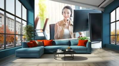 Attractive and smile young asian customer servicer woman wearing headset in modern creative meeting working office sit at computer monitor thump up ,work from and social distancing concept. Wall mural