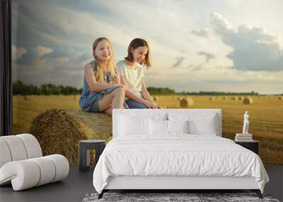 Adorable young sisters having fun in a wheat field on a summer day. Children playing at hay bale field during harvest time. Kids enjoying warm sunset outdoors. Wall mural