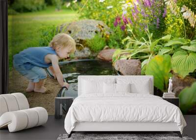 Funny toddler boy having fun by a small garden pond on sunny summer day. Child exploring nature. Wall mural