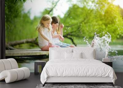 Two cute little girls sitting on a wooden platform by the river or lake dipping their feet in the water on warm summer day Wall mural