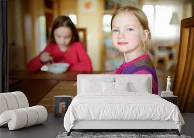 Two cute little sisters eating a soup in cozy dining room. Children having a dinner at home. Wall mural