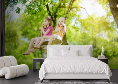 Two cute little sisters having fun on a swing together in beautiful summer garden on warm and sunny day outdoors Wall mural