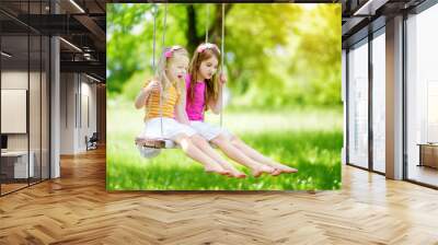 Two cute little sisters having fun on a swing together in beautiful summer garden on warm and sunny day outdoors Wall mural