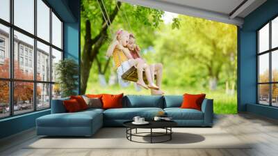 Two cute little sisters having fun on a swing together in beautiful summer garden on warm and sunny day outdoors Wall mural