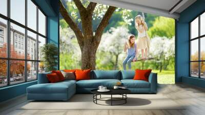 Two cute sisters having fun on a swing in blossoming old apple tree garden outdoors on sunny spring day. Wall mural