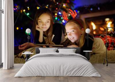 Two cute young sisters using a tablet pc at home by a fireplace in warm and cozy living room on Christmas eve. Winter evening with family and kids. Wall mural