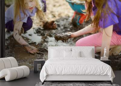 Two funny little girls playing in a large wet mud puddle on sunny summer day. Children getting dirty while digging in muddy soil. Wall mural