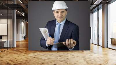 Handsome confident engineer in hard hat with clipboard and blueprint roll looking at camera with smile isolated on grey background. Successful man from industrial sphere Wall mural