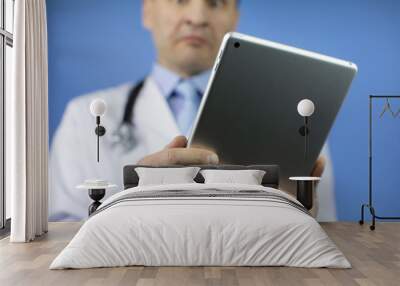 The doctor looks at the tablet with the results of the patient's tests. Adult male physician with stethoscope dressed in white medical coat holding a tablet. Blue background. Wall mural