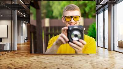 Young happy hipster guy in yellow T-shirt and trendy sunglasses have fun outdoors, takes photo with instant camera on green nature background, resting time. Summer, leisure, photography hobby concept Wall mural