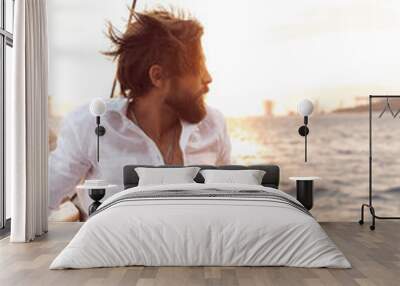 handsome man with a beard at sunset on a yacht wearing a white linen shirt and pants with a brutal expression Wall mural