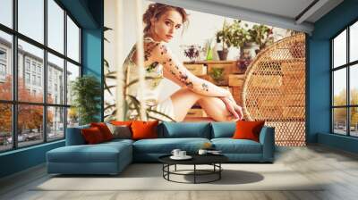 Portrait of beautiful young sexy woman with updo hairstyle and flower tattoos on her arms and shoulders wearing silk floral dress in studio with plants and straw chair Wall mural