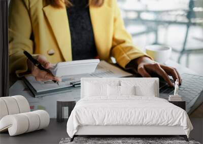 A businesswoman in a yellow blazer uses a calculator and laptop to analyze financial documents in a modern office environment. Wall mural