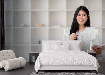 A woman is standing in front of a white wall with a book in her hand. She is smiling and she is happy Wall mural