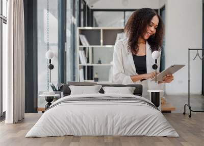 Focused entrepreneur in a white blazer reviews data on her tablet in a modern workspace with a laptop on the desk. Wall mural