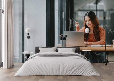 Introspective businesswoman contemplates her work, pen in hand, while sitting at her laptop in a well-organized office space. Wall mural