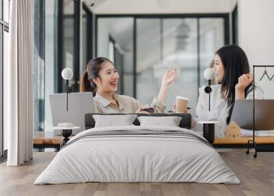 Two cheerful women having a coffee break with laptops open on their office table, engaging in a lively conversation. Wall mural