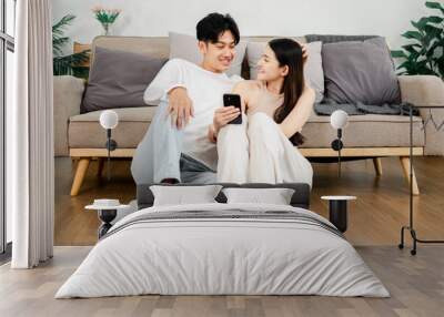 Young Asian couple sits cozily on a beige sofa, sharing a moment with a smartphone in a well-lit, modern living room. Wall mural