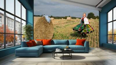 Blonde pretty young smiling girl dressed in Polish national folk dress dancing and spinning around in the field of harvest straw bales Wall mural