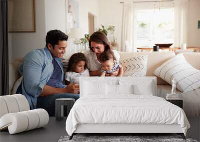 Hispanic couple sitting on the sofa reading a book at home with their baby son and young daughter Wall mural