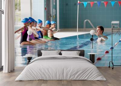 Male Coach In Water Gives Children Instructions In Swimming Lesson As They Sit On Edge Of Pool Wall mural