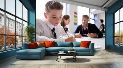 Male High School Student Wearing Uniform Working At Table With Teacher Talking To Pupils In Background Wall mural