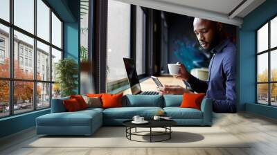 Mid adult black male creative sits by window having coffee, using a laptop and smartphone, side view Wall mural