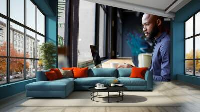 Mid adult black male creative sitting by window in an office social area using a laptop, side view Wall mural