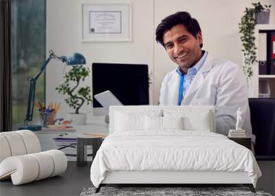 Portrait Of Male Doctor Or GP Wearing White Coat Sitting At Desk In Office Using Digital Tablet Wall mural