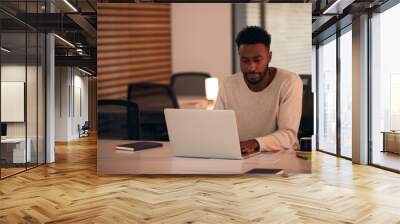 Young Businessman Working Late Sitting At Desk With Laptop In Modern Open Plan Office Wall mural