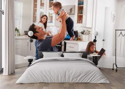 Young Hispanic family in their kitchen, dad lifting baby in the air, mum cooking at hob, close up Wall mural