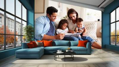 Young Hispanic family of four sitting on the sofa reading a book together in their living room Wall mural