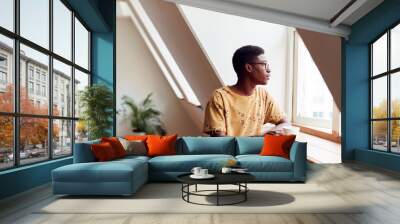 Young Man Relaxing In Loft Apartment Looking Out Of Window With Hot Drink Wall mural